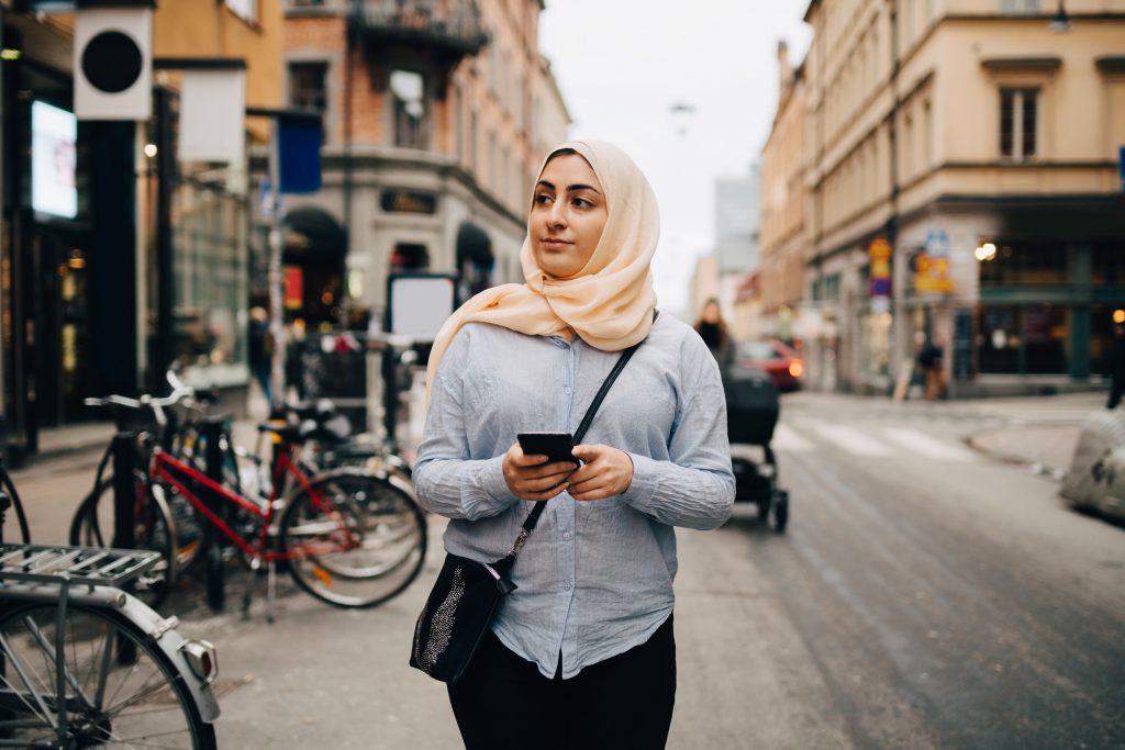 Young Muslim woman looking away holding smart phone while walking on street in city